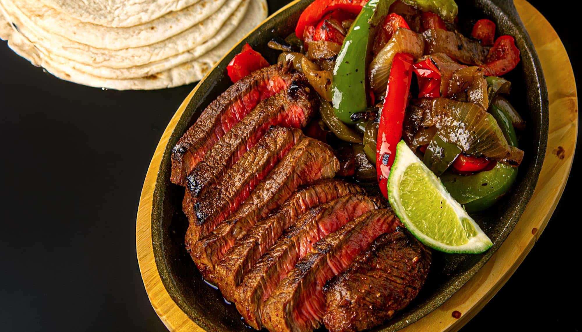 Grilled sliced steak with sautéed bell peppers and onions, garnished with a lime wedge, served with warm tortillas on the side.