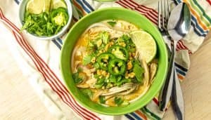 Thai chicken coconut soup with rice noodles, jalapeño, cilantro, peanuts, and lime wedge in a green bowl with extra herbs on the side.