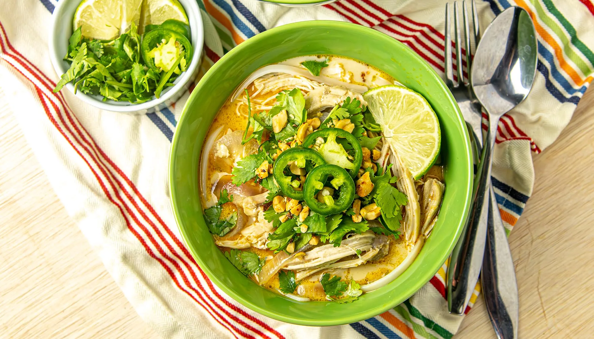 Thai chicken coconut soup with rice noodles, jalapeño, cilantro, peanuts, and lime wedge in a green bowl with extra herbs on the side.