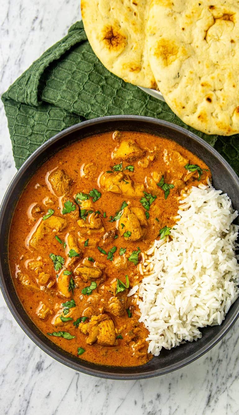 Chicken curry garnished with cilantro, served with white rice and naan bread on a green cloth over a marble surface.