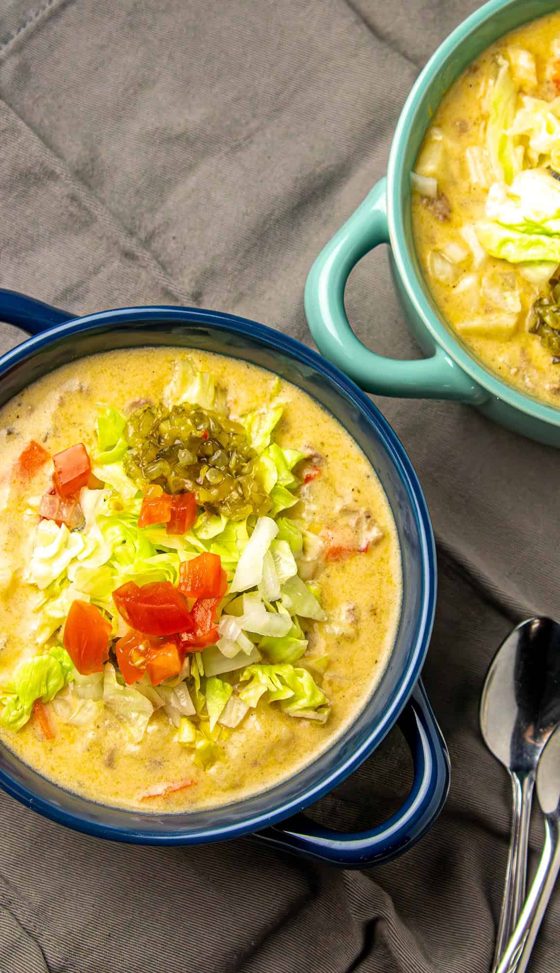 Creamy soup topped with chopped lettuce, diced tomatoes, onions, and relish in blue and teal bowls on gray cloth with two spoons nearby.