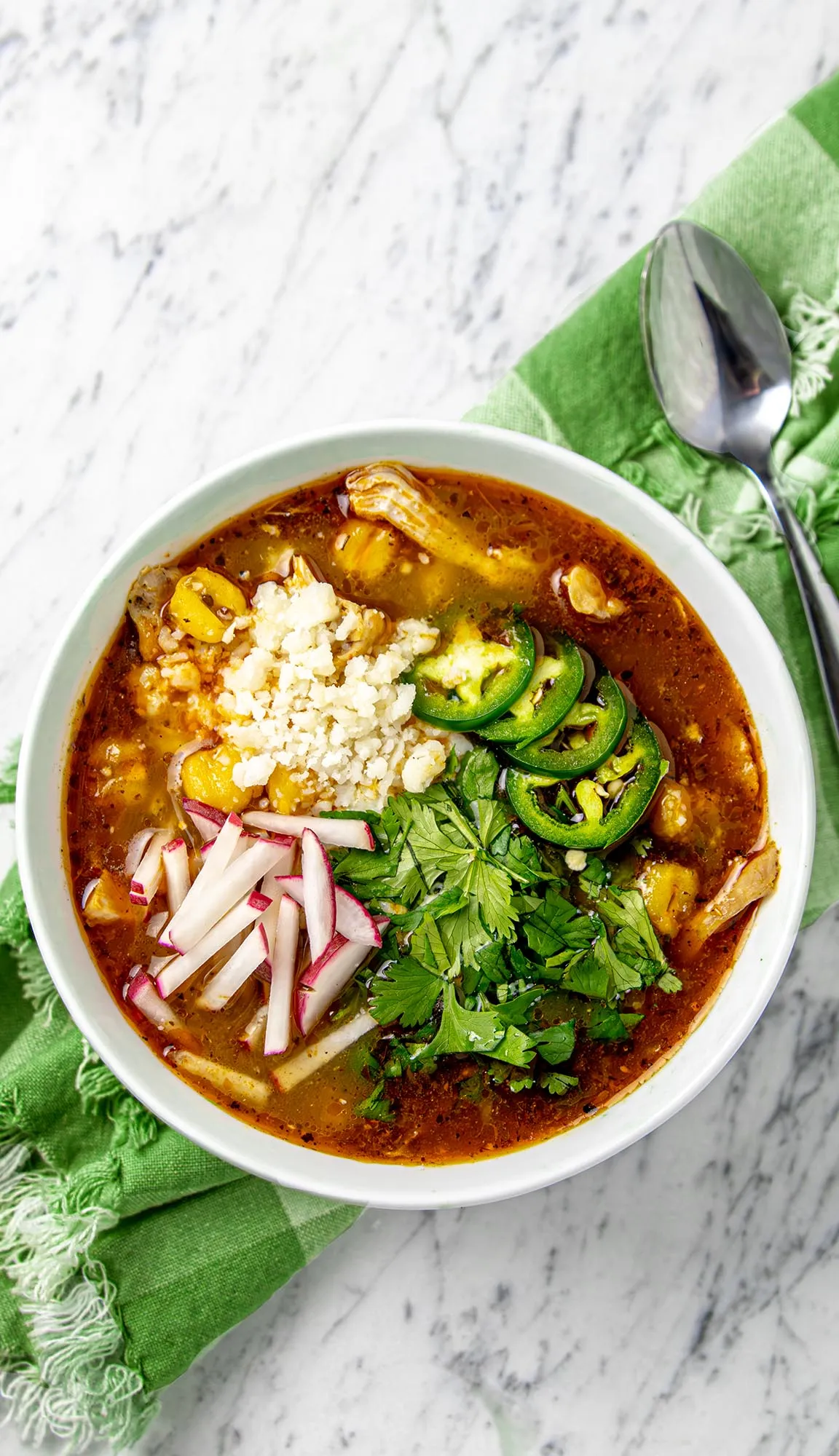 Bowl of chicken pozole with hominy, topped with sliced radishes, jalapeños, cilantro, and crumbled cheese, on a green cloth with a spoon.
