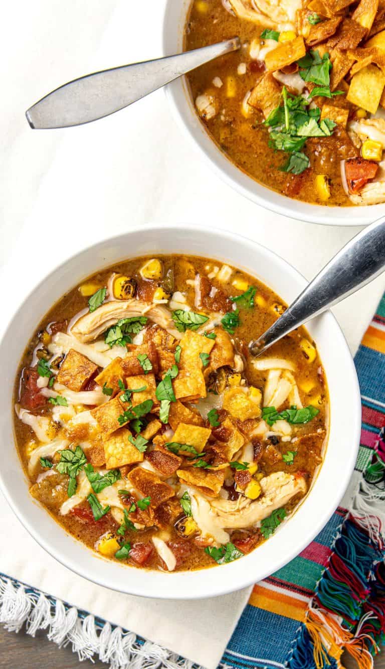 Bowls of chicken tortilla soup topped with tortilla strips, shredded cheese, corn, diced tomatoes, and chopped cilantro, with spoons inside.