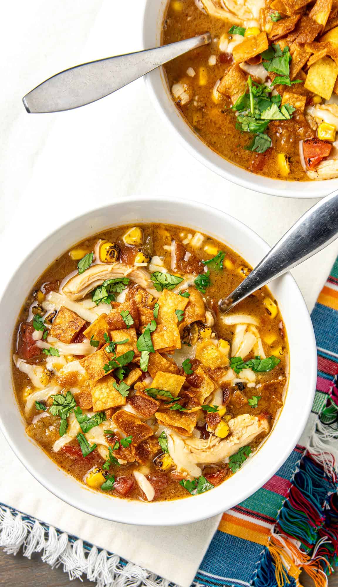 Bowls of chicken tortilla soup topped with tortilla strips, shredded cheese, corn, diced tomatoes, and chopped cilantro, with spoons inside.