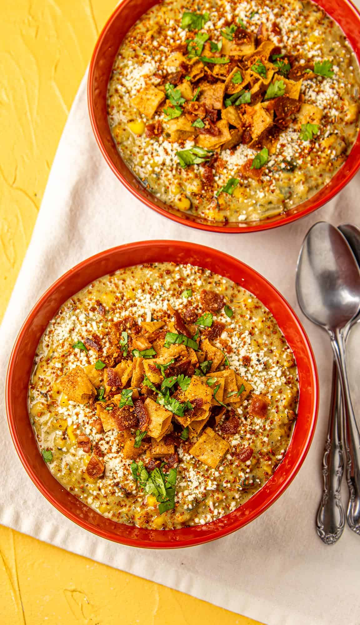 Two red bowls of creamy chili topped with crushed chips, bacon, cheese, and cilantro on a beige cloth with two spoons beside them.