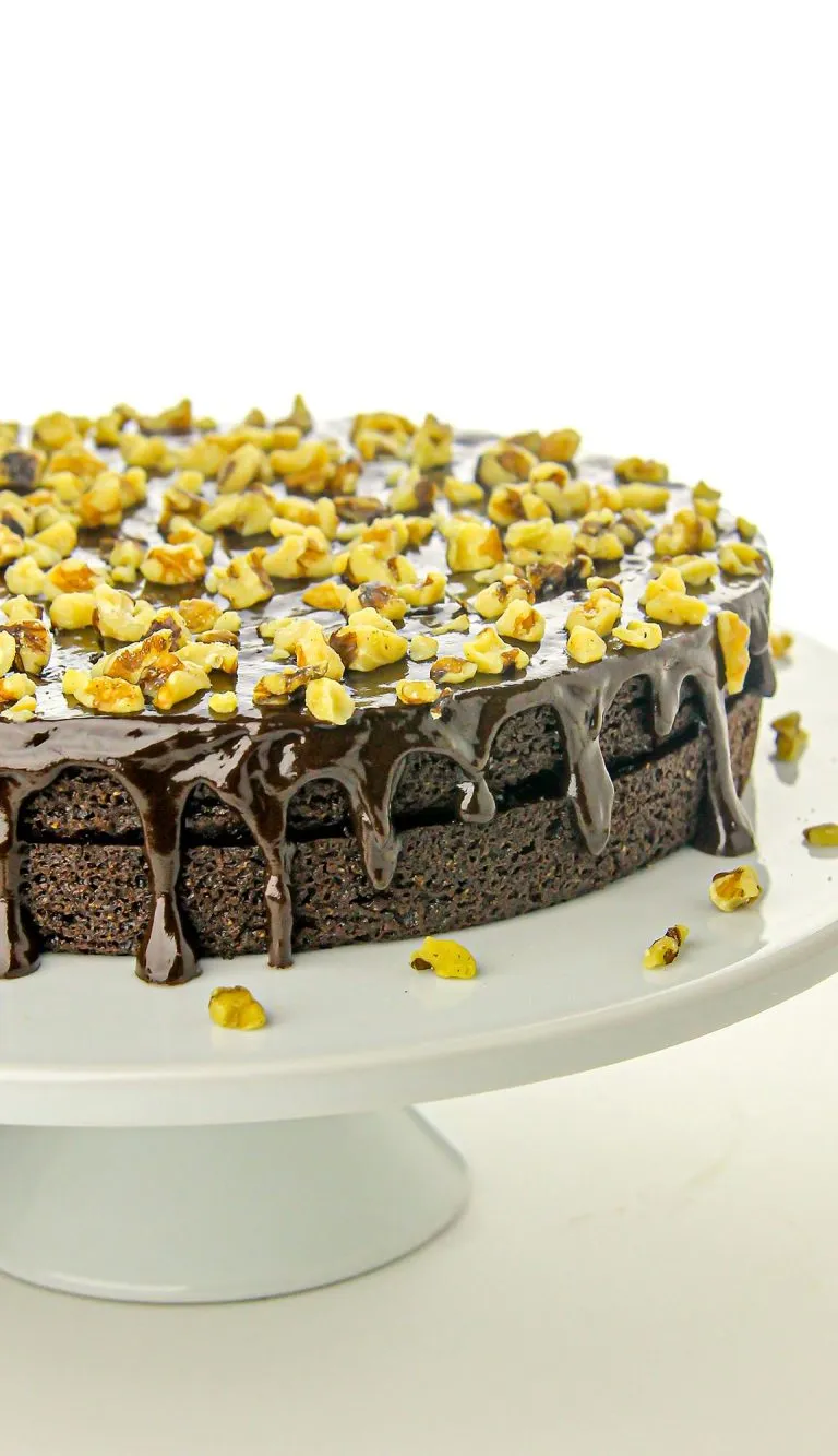 Chocolate cake with glossy ganache dripping over the edges, topped with chopped walnuts on a white cake stand.