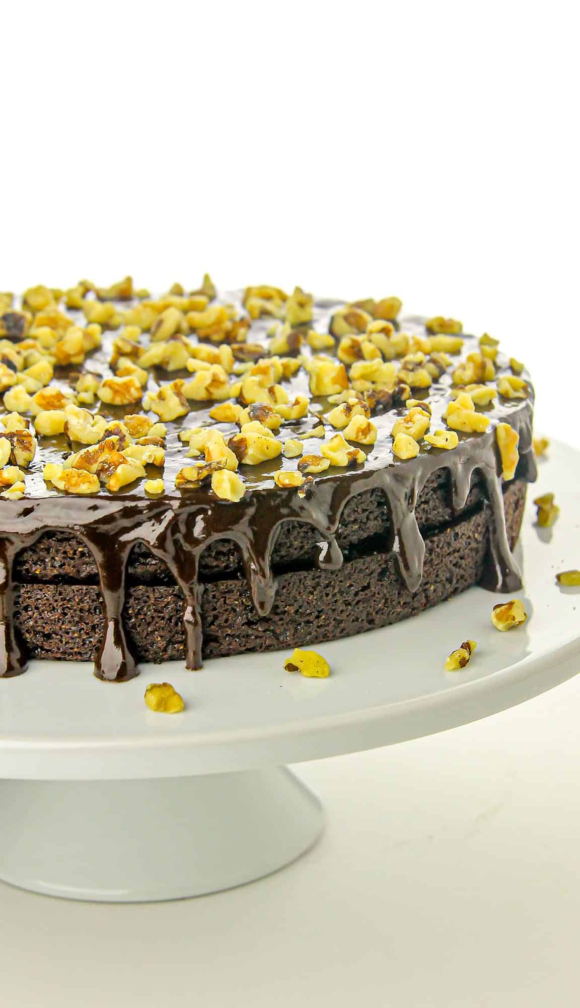 Chocolate cake with glossy ganache dripping over the edges, topped with chopped walnuts on a white cake stand.