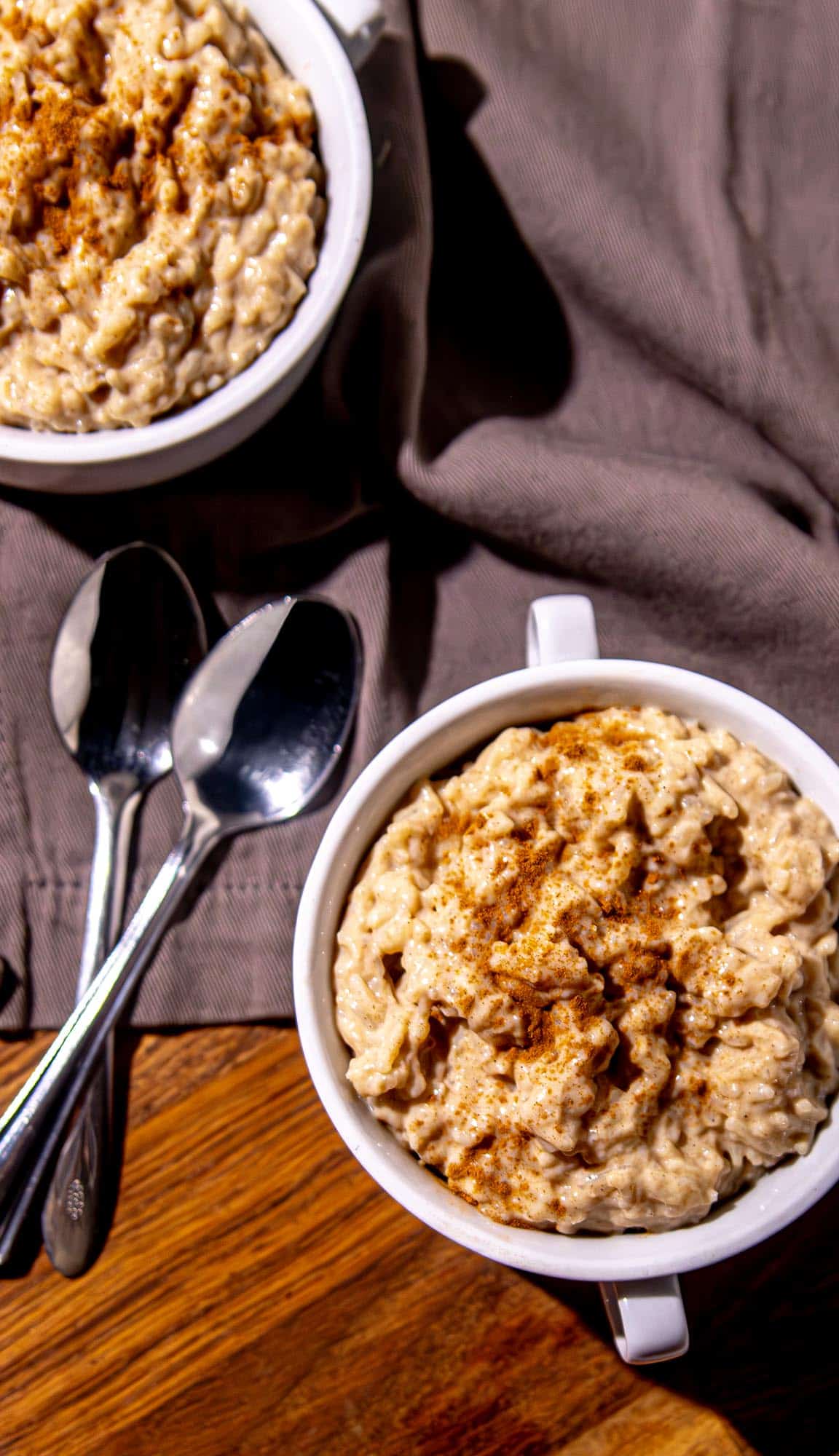 Two white mugs filled with creamy oatmeal sprinkled with cinnamon, placed on a brown cloth with two metal spoons beside them.