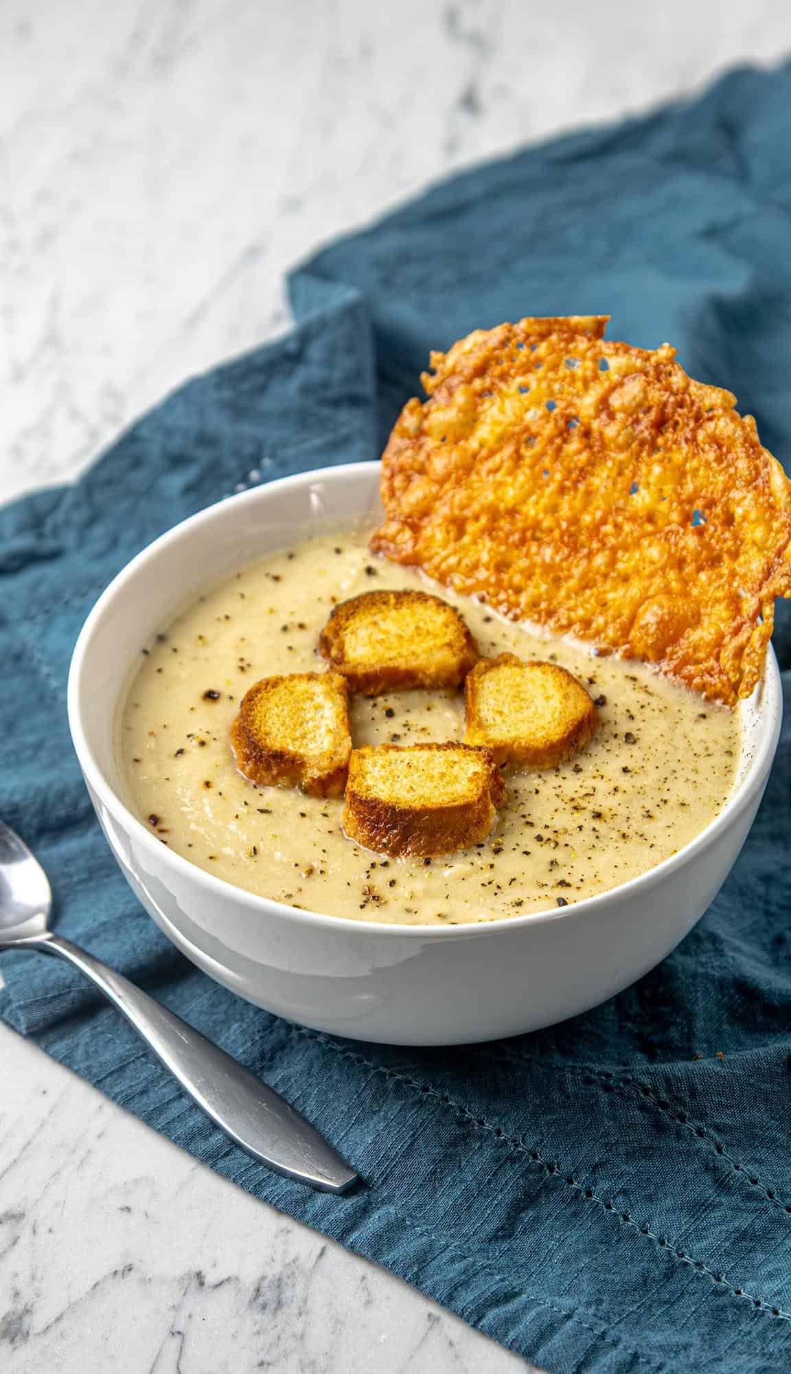 Creamy soup garnished with black pepper, toasted bread cubes, and a large crispy cheese crisp on a blue napkin with a spoon nearby.