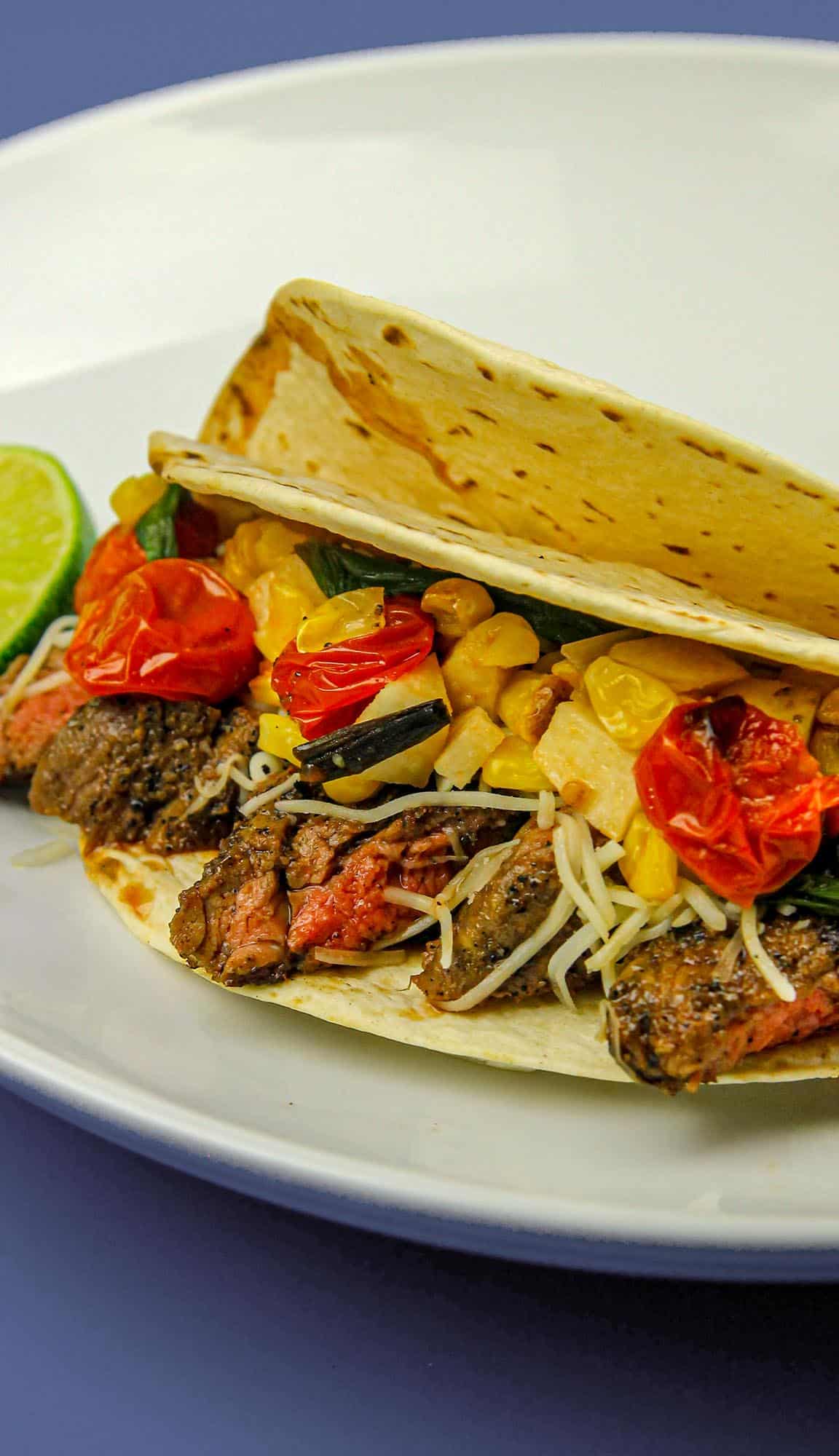 Steak taco with grilled cherry tomatoes, corn, cheese, and greens in a soft tortilla, served with a lime wedge on a white plate.