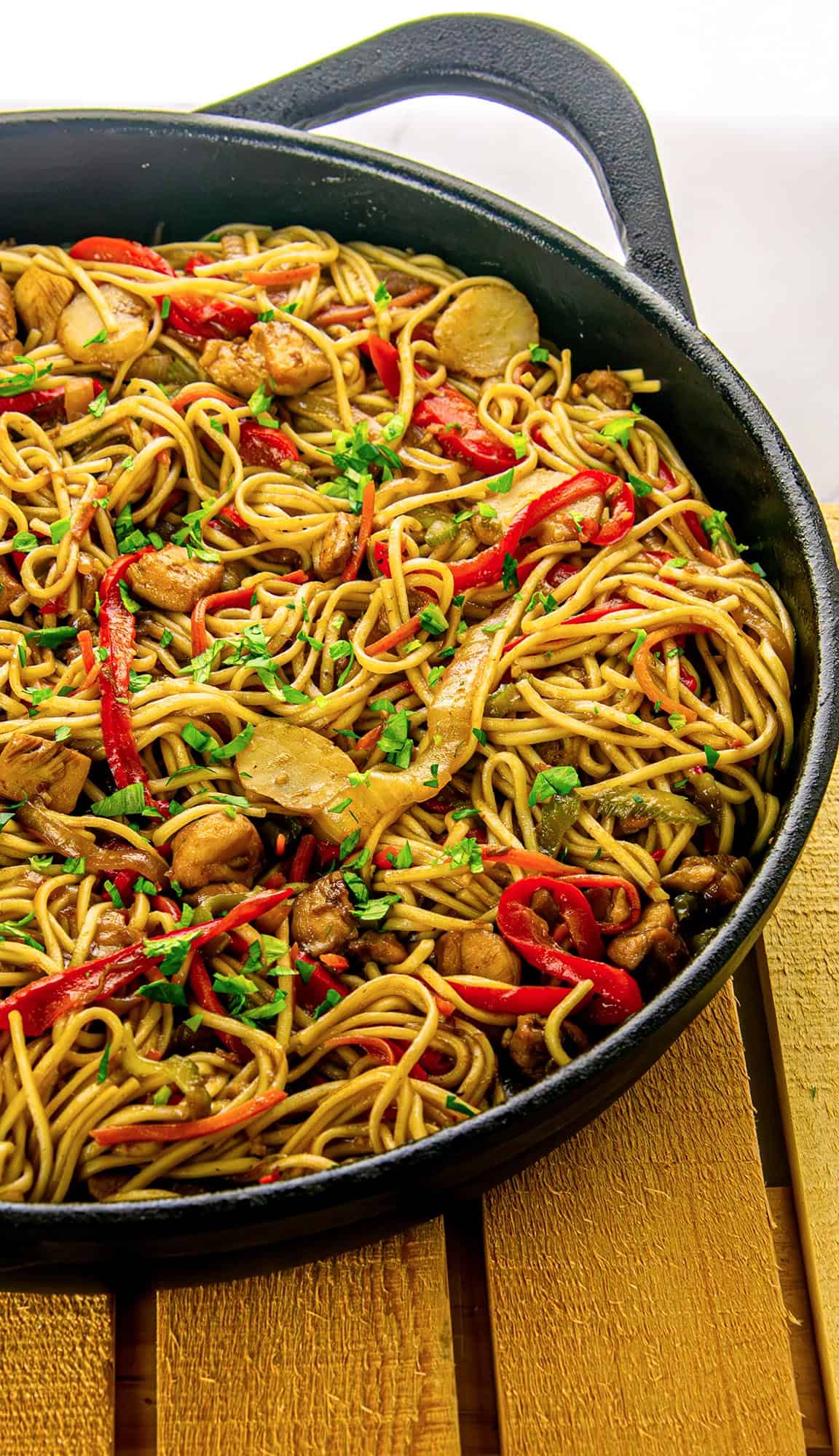 Stir-fried noodles with chicken, red bell peppers, mushrooms, and garnished with chopped green herbs in a black cast iron pan.