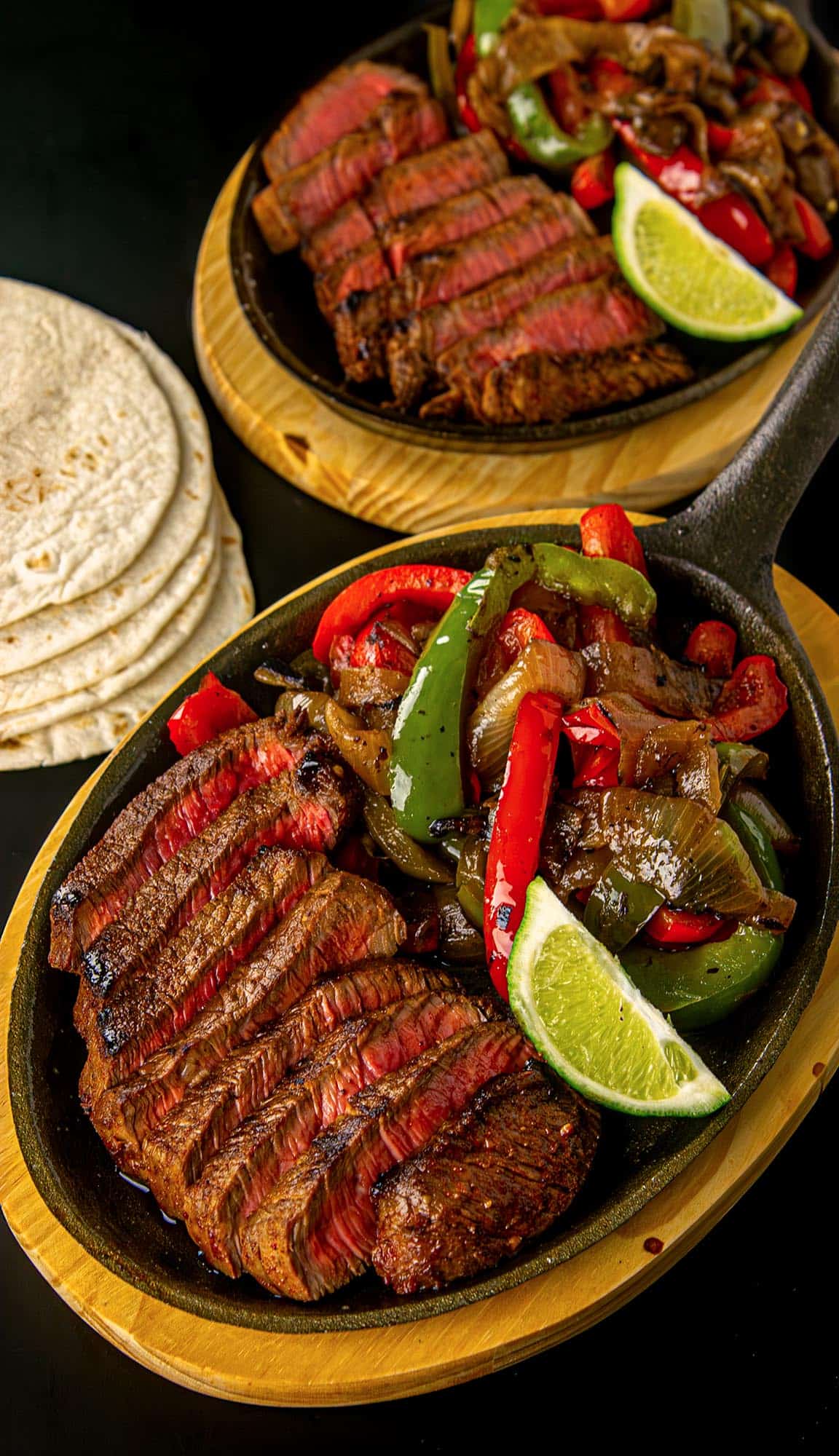 Sliced medium-rare steak with sautéed green and red bell peppers and onions, lime wedge, and a stack of flour tortillas on the side.