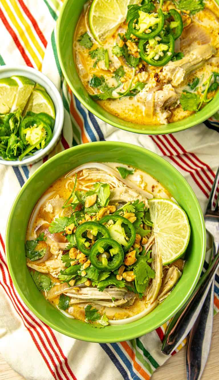 Two bowls of chicken coconut soup with lime wedges, cilantro, jalapeño slices, chopped peanuts, and a small bowl of lime and jalapeño on a striped cloth.