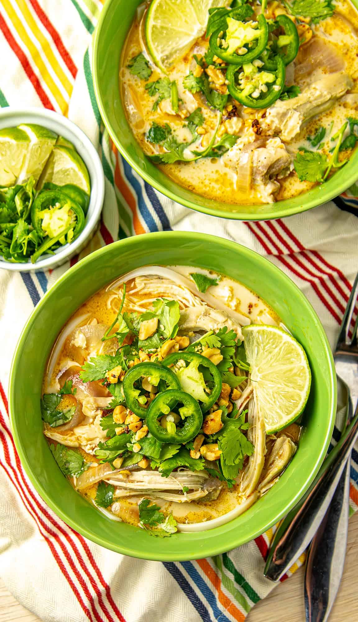 Two bowls of chicken coconut soup with lime wedges, cilantro, jalapeño slices, chopped peanuts, and a small bowl of lime and jalapeño on a striped cloth.