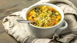 White bowl of creamy soup with pasta, sausage, spinach, and herbs, placed on a beige cloth on a wooden surface.