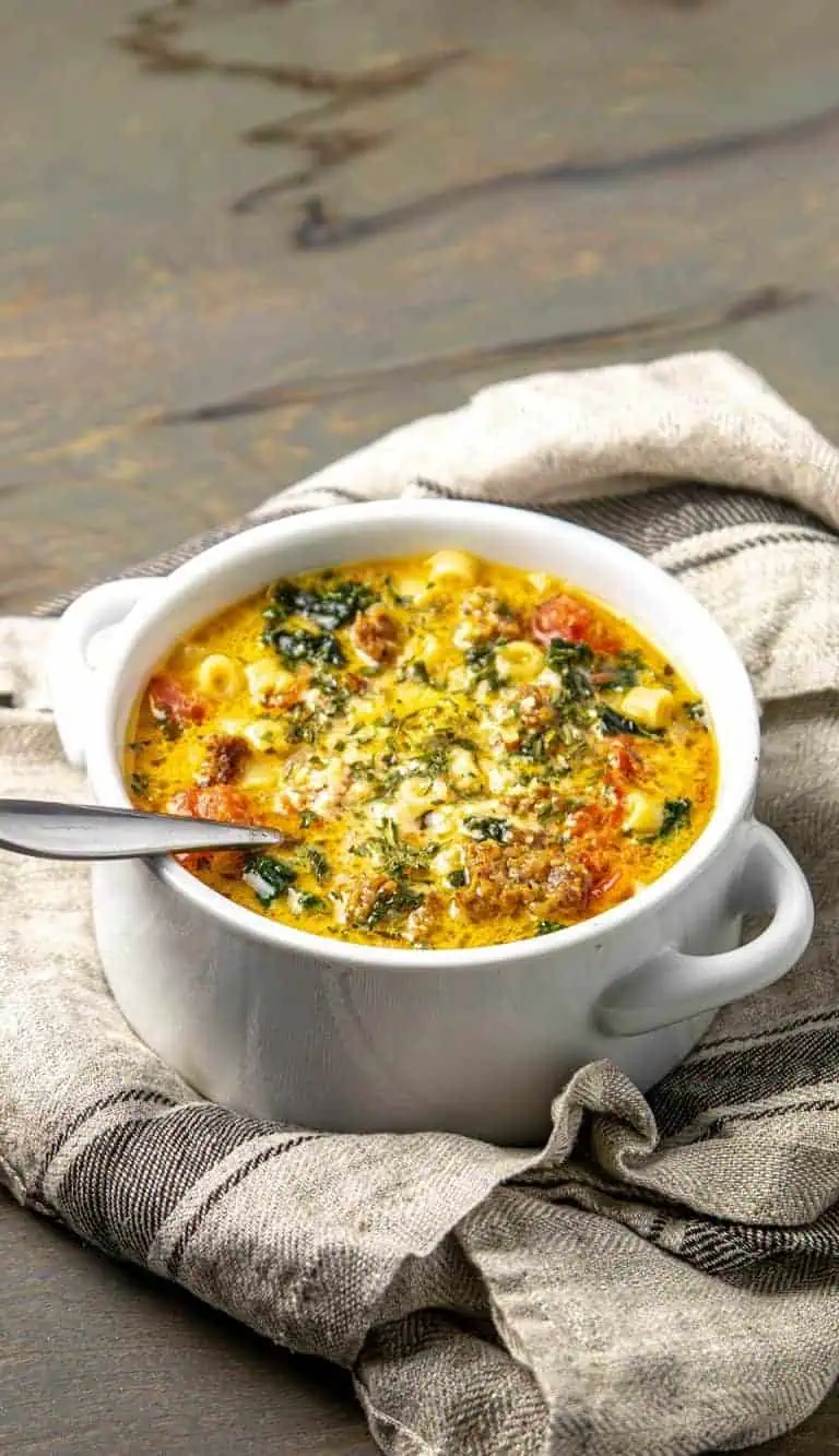 Creamy soup with sausage, macaroni, spinach, and herbs in a white bowl on a textured cloth and wooden surface.