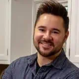 Smiling man with short dark hair and beard wearing a dark blue button-up shirt in a kitchen setting.