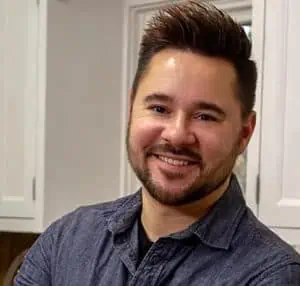 Smiling man with short dark hair and beard wearing a dark blue button-up shirt in a kitchen setting.