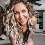 Smiling woman with long curly blonde hair and nose ring leaning forward in a bedroom with shelves and hanging clothes in the background.