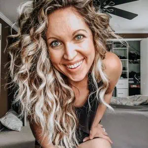Smiling woman with long curly blonde hair and nose ring leaning forward in a bedroom with shelves and hanging clothes in the background.