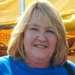 Middle-aged woman with blonde hair wearing a blue shirt, smiling outdoors under a yellow canopy.