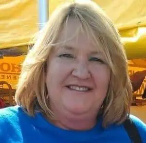 Middle-aged woman with blonde hair wearing a blue shirt, smiling outdoors under a yellow canopy.