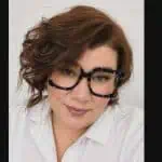 Woman with wavy brown hair, wearing large tortoiseshell glasses, a white collared shirt, and light pink lipstick, smiling softly.