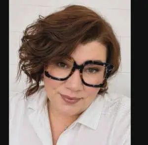 Woman with wavy brown hair, wearing large tortoiseshell glasses, a white collared shirt, and light pink lipstick, smiling softly.