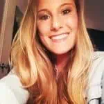 Young woman with long blonde hair smiling, wearing a light denim jacket, indoors near a door and a dark background.