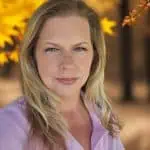 Blonde woman with light green eyes wearing a light pink shirt, with blurred autumn leaves in yellow and orange in the background.