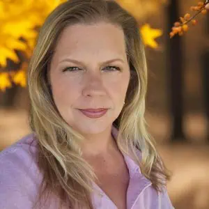 Blonde woman with light green eyes wearing a light pink shirt, with blurred autumn leaves in yellow and orange in the background.