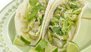 Two fish tacos with lettuce, cilantro, sauce, and lime wedges on a light green plate.