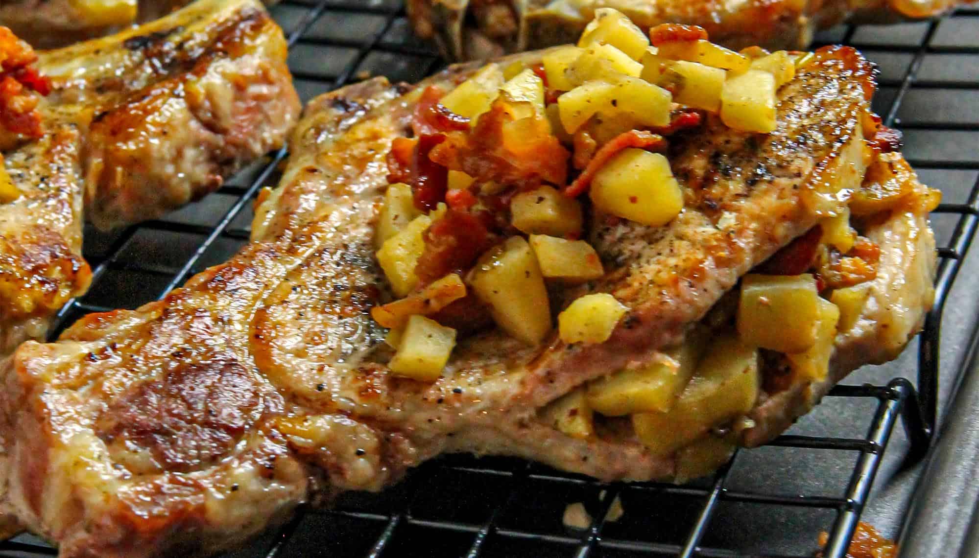 Grilled pork chops topped with cooked diced apples and bacon on a cooling rack.