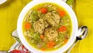 Chicken and vegetable soup with three dumplings in a white bowl on a yellow placemat next to a spoon.
