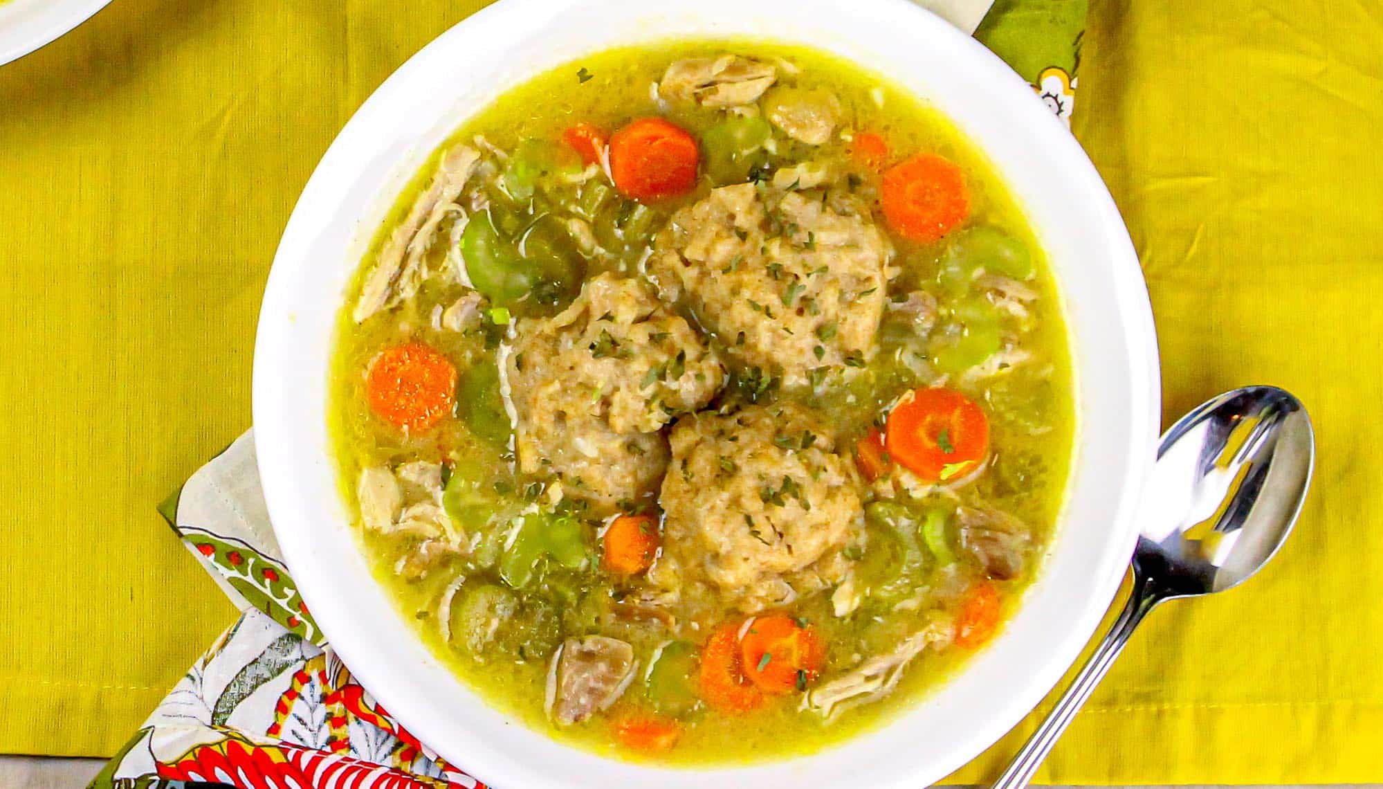 Chicken and vegetable soup with three dumplings in a white bowl on a yellow placemat next to a spoon.