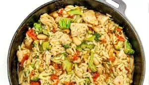Cast iron skillet of cooked chicken, broccoli, white beans, orzo pasta, and tomato pieces in broth.