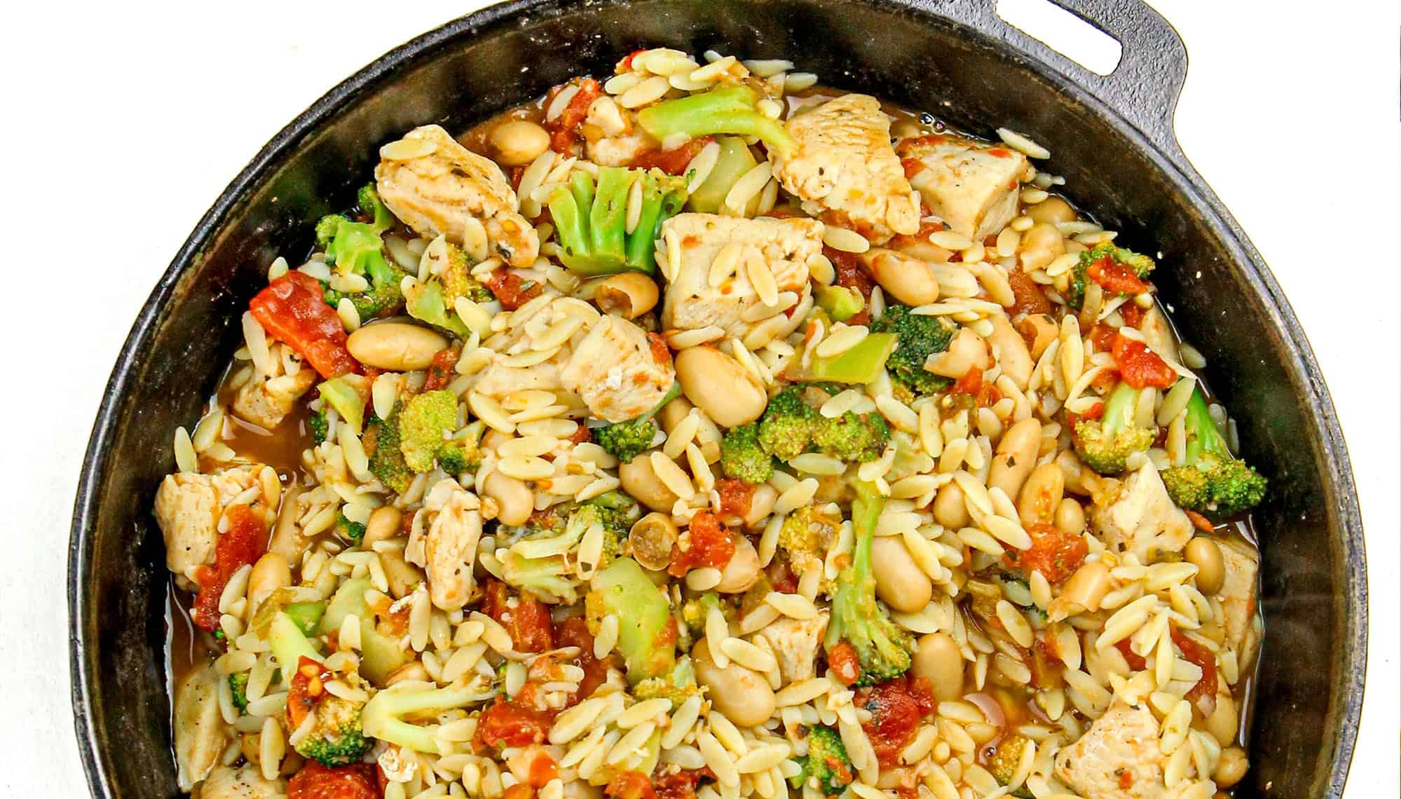 Cast iron skillet of cooked chicken, broccoli, white beans, orzo pasta, and tomato pieces in broth.