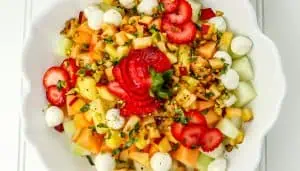 Colorful fruit salad with strawberries, cantaloupe, honeydew, mozzarella balls, chopped nuts, and herbs in a white bowl.