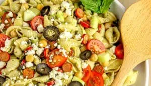 Pasta salad with cherry tomatoes, black olives, chickpeas, cucumber, feta cheese, and basil leaves in a white bowl with a wooden spoon.