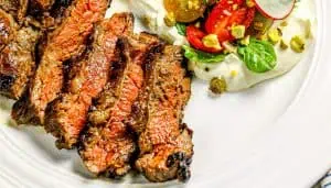 Grilled sliced steak with a side of whipped ricotta topped with cherry tomatoes, spinach, radish, and chopped pistachios on a white plate.