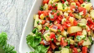 Colorful pineapple salsa with red tomatoes, green peppers, red onions, and cilantro in a white bowl on a wooden surface.