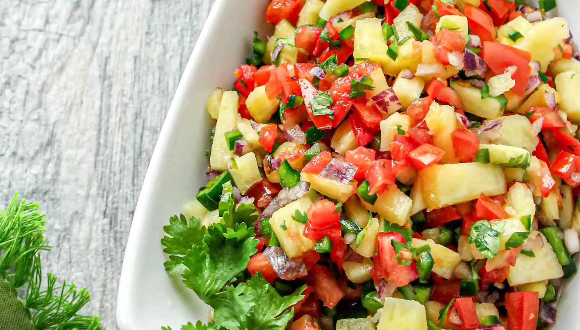 Colorful pineapple salsa with red tomatoes, green peppers, red onions, and cilantro in a white bowl on a wooden surface.
