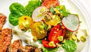 Grilled steak slices with whipped ricotta cheese, topped with spinach, cherry tomatoes, radish slices, and crushed pistachios on a white plate.
