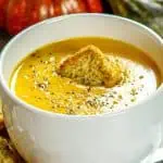 Creamy butternut squash soup in a white mug, garnished with cracked black pepper and a triangular crouton, with additional croutons on the side.