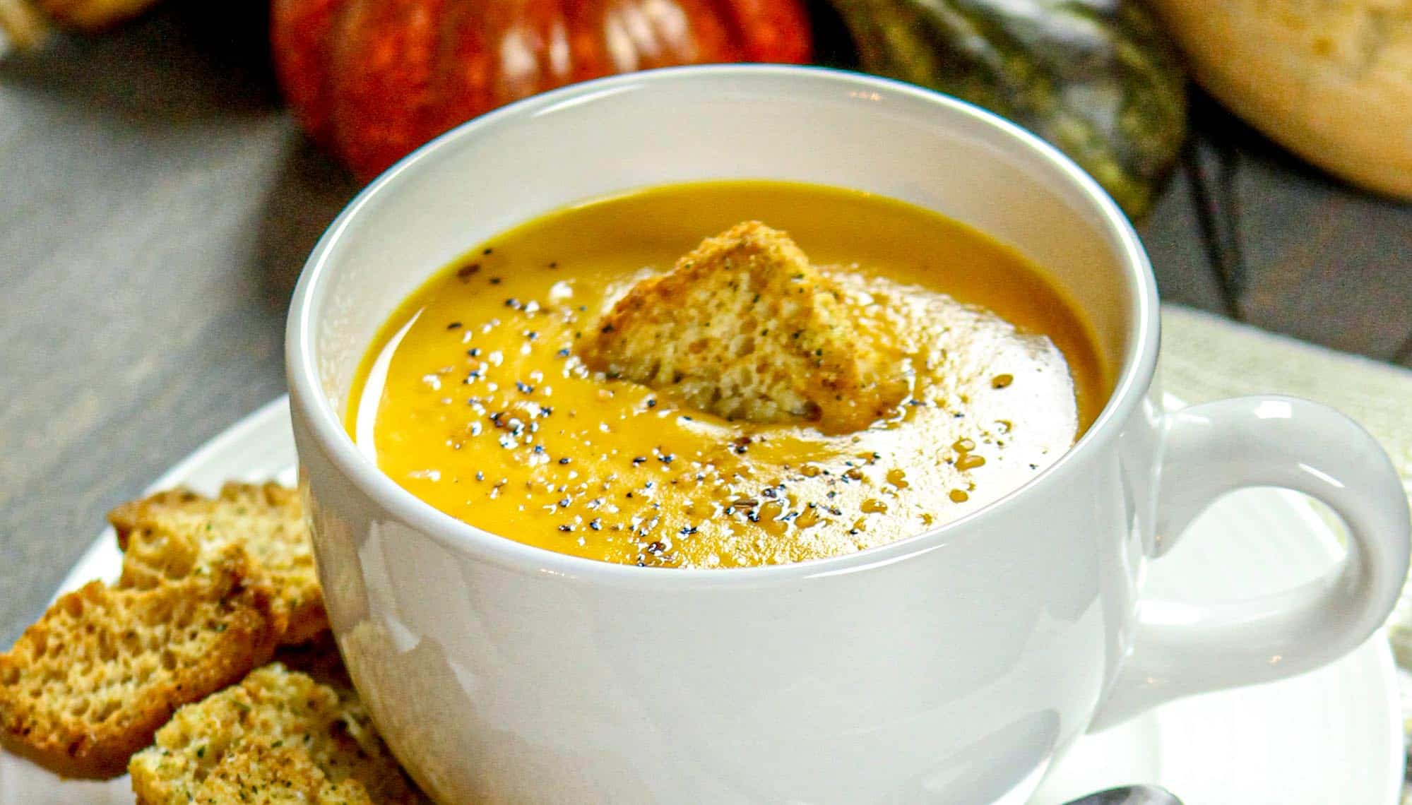Creamy butternut squash soup in a white mug, garnished with cracked black pepper and a triangular crouton, with additional croutons on the side.