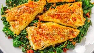 Three seasoned and cooked fish fillets served on sautéed spinach with small bits of tomato on a white plate.