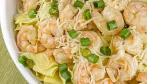 Shrimp pasta with shredded cheese and chopped green onions in a white bowl on a green surface.