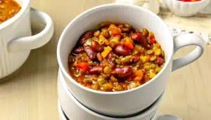 White cup filled with lentil and kidney bean vegetable chili on a light wooden surface.