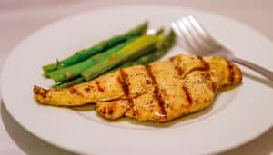Grilled chicken breast with visible grill marks served with steamed asparagus on a white plate with a fork.