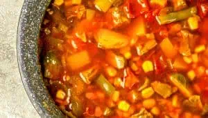 Chunky vegetable soup with corn, green beans, potatoes, tomatoes, and broth in a black speckled pot.